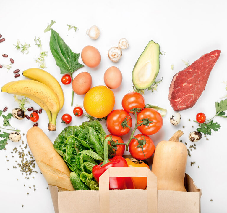 A paper bag filled with fresh vegetables, fruits, bread, and eggs is surrounded by assorted foods—each showcasing supply chain traceability—including bananas, tomatoes, spinach, steak, avocado, oranges, mushrooms, and quail eggs on a white background.
