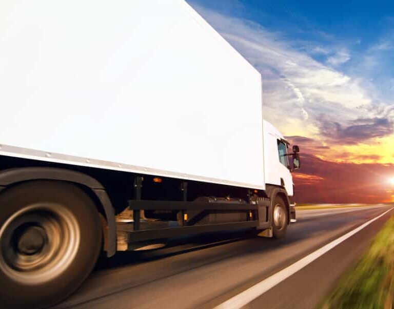 A white delivery truck speeds down a highway at sunset, framed by dramatic blue, orange, and yellow skies—a striking scene that captures the efficiency powered by modern transportation software.