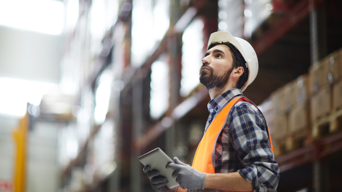 A warehouse worker wearing a white hard hat, orange safety vest, and gloves holds a tablet while looking up at shelves stocked with boxes in a large, brightly lit warehouse, supporting efficient master planning.