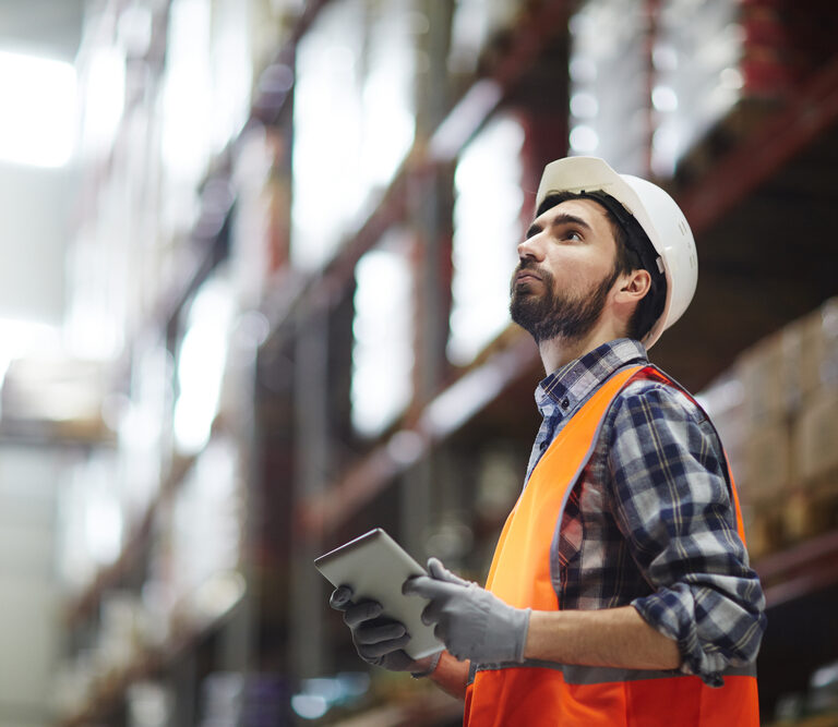 A warehouse worker wearing a white hard hat, orange safety vest, and gloves holds a tablet while looking up at shelves stocked with boxes in a large, brightly lit warehouse, supporting efficient master planning.