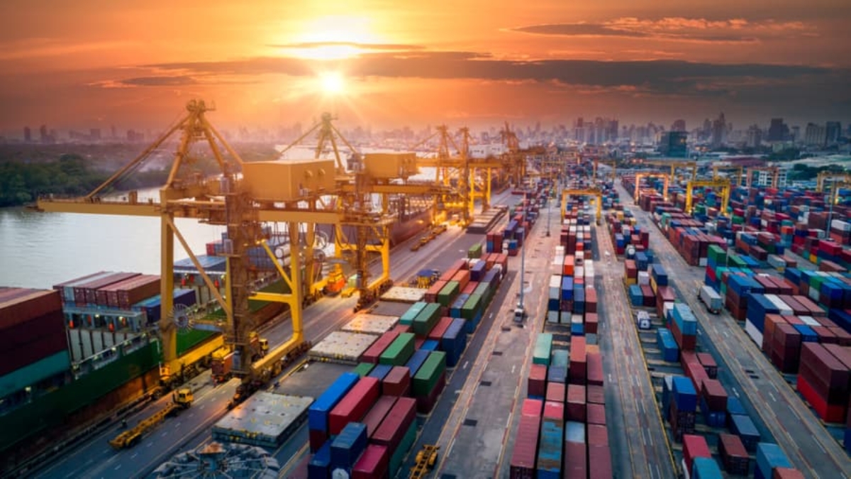 A busy shipping port at sunset with large cranes, colorful stacked shipping containers, and a city skyline in the background.