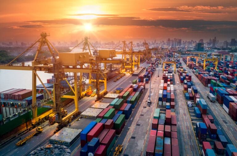 A busy shipping port at sunset with large cranes, colorful stacked shipping containers, and a city skyline in the background.