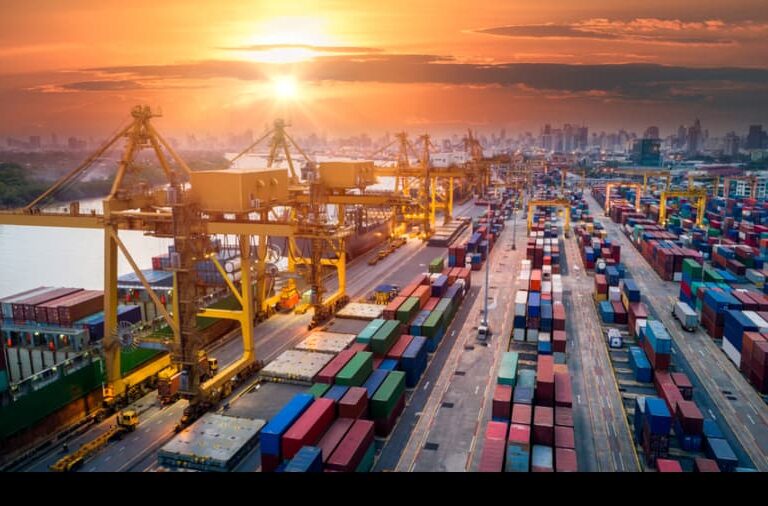 A busy shipping port at sunset with large cranes loading and unloading colorful shipping containers; city skyline is visible in the background.
