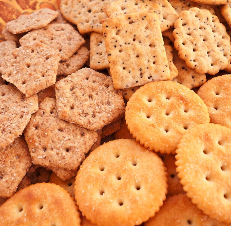 An assortment of different crackers, including hexagonal wheat crackers, square seeded crackers, and round buttery crackers, arranged close together on a patterned surface.