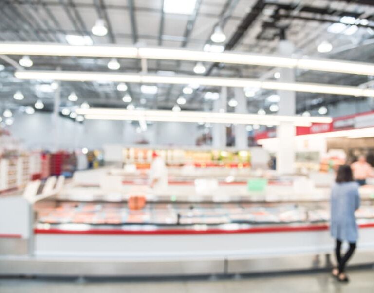 A blurred image of a supermarket interior with bright overhead lights, aisles, and display cases. A person stands near the counter in the foreground, but all details are out of focus.