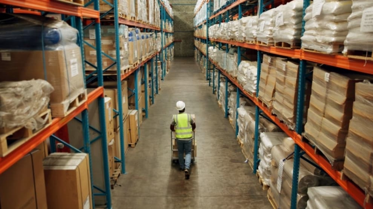 A worker in a high-visibility vest and hard hat pushes a cart down a wide aisle in the Blue Ridge warehouse, surrounded by tall shelves filled with various boxes and packages.