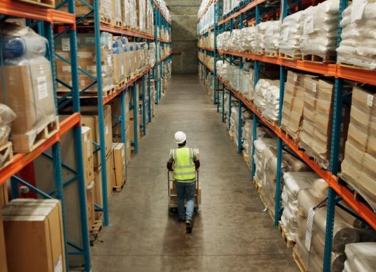 A worker in a high-visibility vest and hard hat pushes a cart down a wide aisle in the Blue Ridge warehouse, surrounded by tall shelves filled with various boxes and packages.