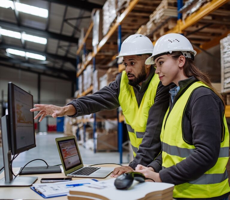 Two warehouse workers in safety vests and hard hats review order accuracy on a computer monitor in a storage facility, surrounded by shelves with stacked boxes and paperwork on a desk.