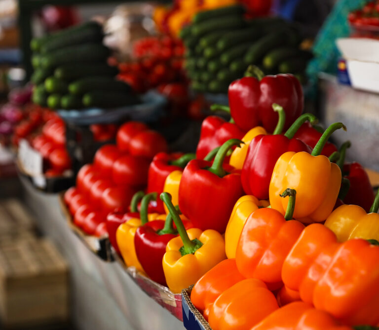 Colorful bell peppers—orange, yellow, and red—are displayed in rows at a market stall, with cucumbers and cherry tomatoes visible in the background, offering a vibrant scene reminiscent of cloud computing’s seamless organization.