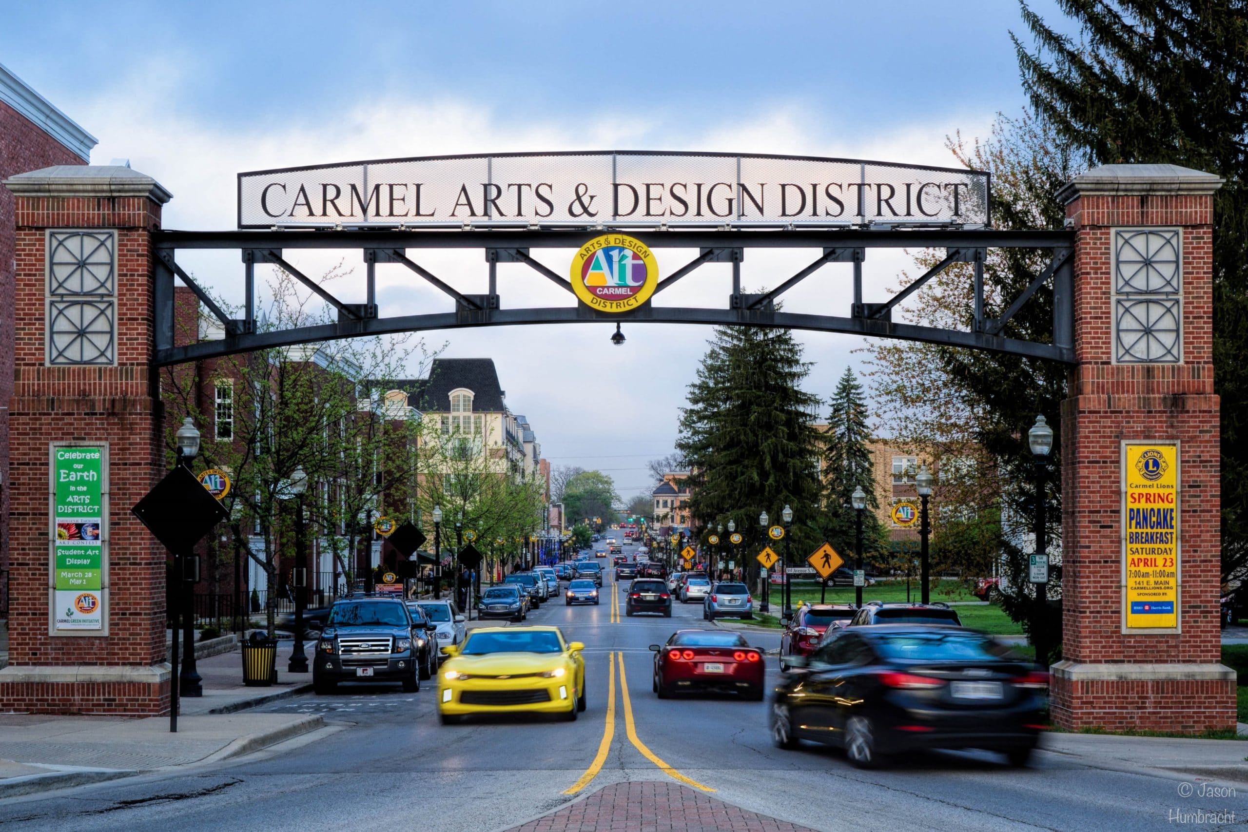 A street scene in the Carmel Arts & Design District shows cars driving under a large decorative archway sign, near shops and buildings lining the street—a vibrant area where enVista Careers are celebrated on a cloudy day.