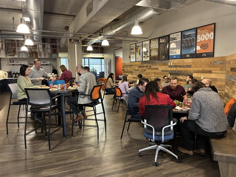 People are gathered in a modern office break room, sitting and standing at tables while eating and talking. Award plaques on the wall showcase achievements, including excellence in parcel audit service and enVista Careers milestones.