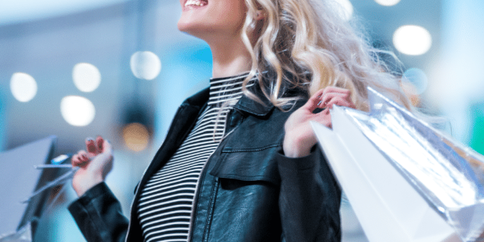 A smiling woman with long blonde hair, wearing a black leather jacket and striped shirt, holds several shopping bags in both hands. The background is blurred with bright lights, suggesting a mall or shopping center.