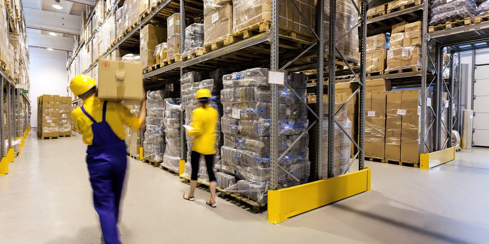 Two workers in safety helmets and uniforms move boxes in a large warehouse with tall shelves stacked with boxes and pallets. The warehouse is brightly lit and organized, with wide aisles for easy movement.