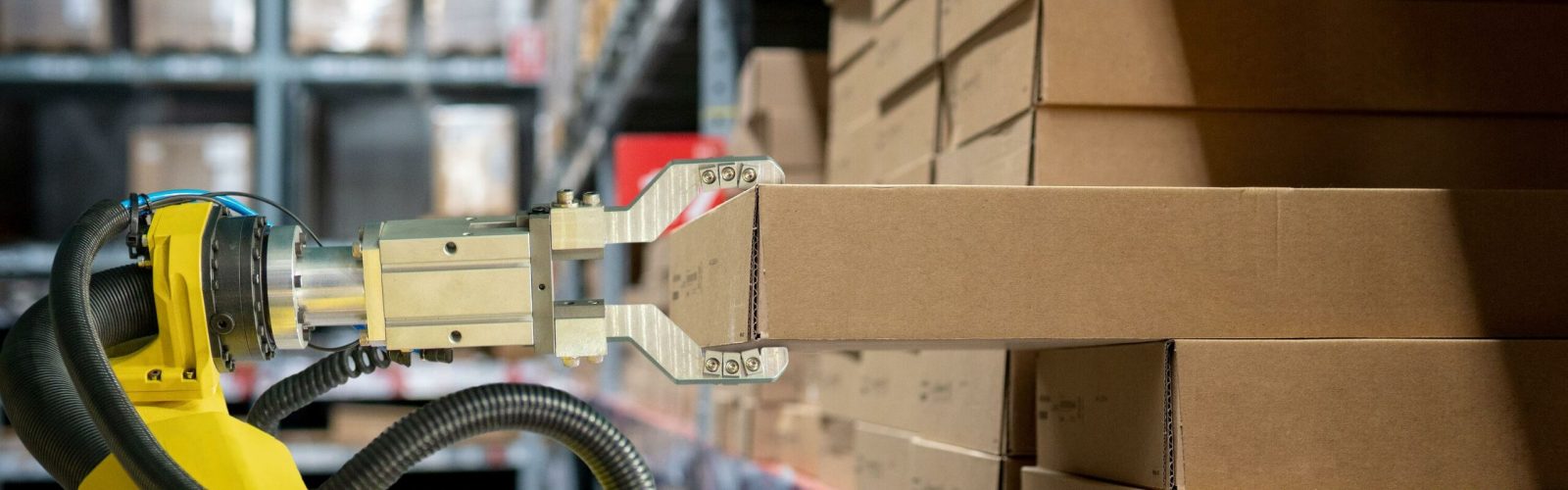 A robotic arm with a metal gripper is picking up a cardboard box from a shelf in a warehouse filled with stacked brown boxes.