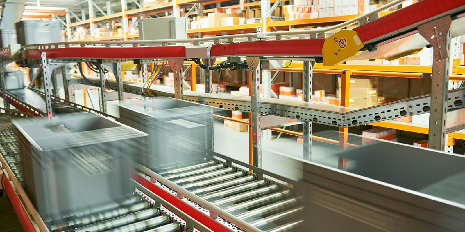 Grey storage bins move quickly along a conveyor belt system in a warehouse, with shelves stocked with various boxes and packages in the background. The motion blur highlights fast-paced automated sorting and supports efficient automation analysis.