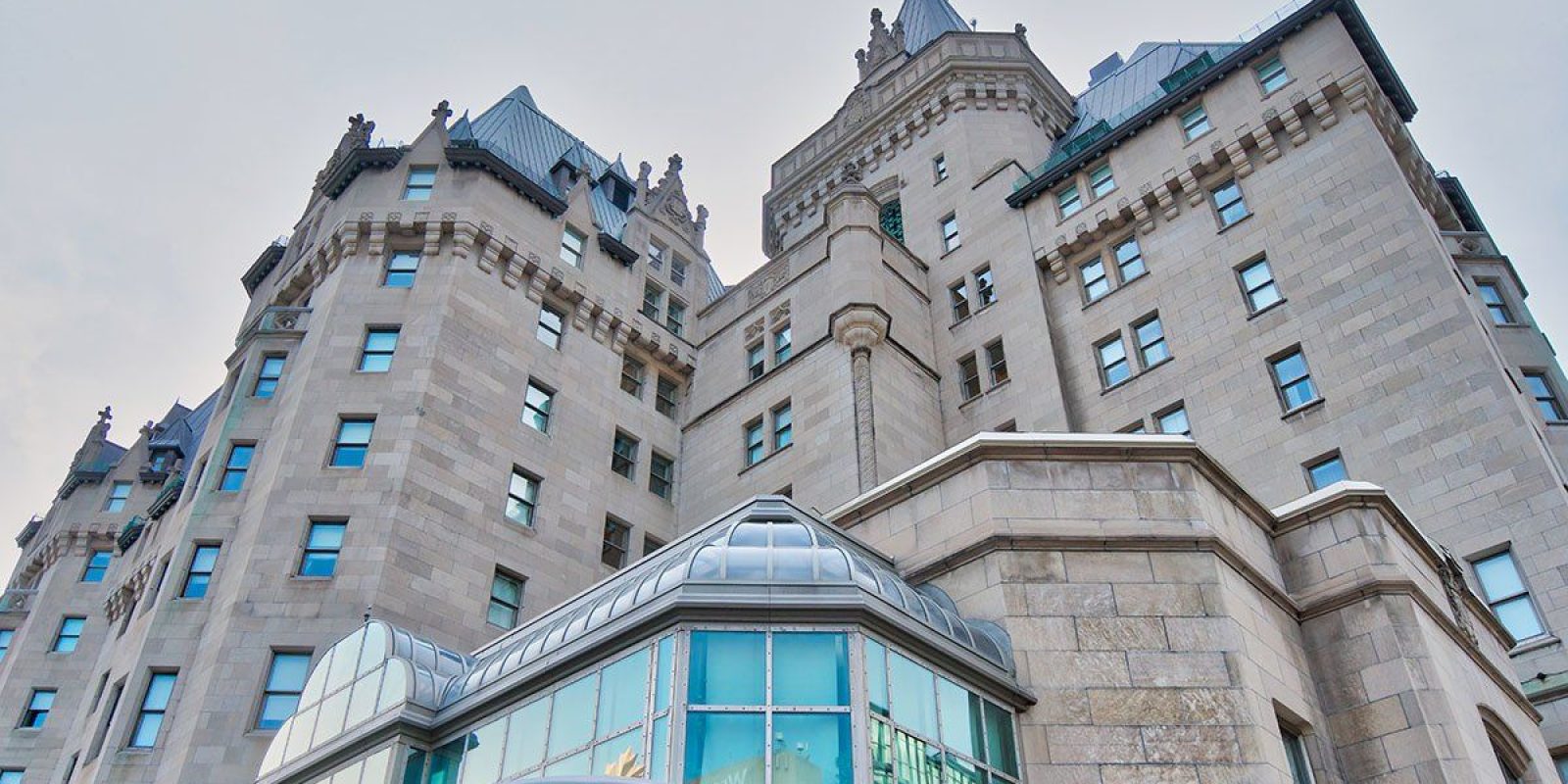 A historic stone building with turrets and steeply pitched roofs, featuring large windows and intricate architectural details, viewed from a low angle against a pale sky.