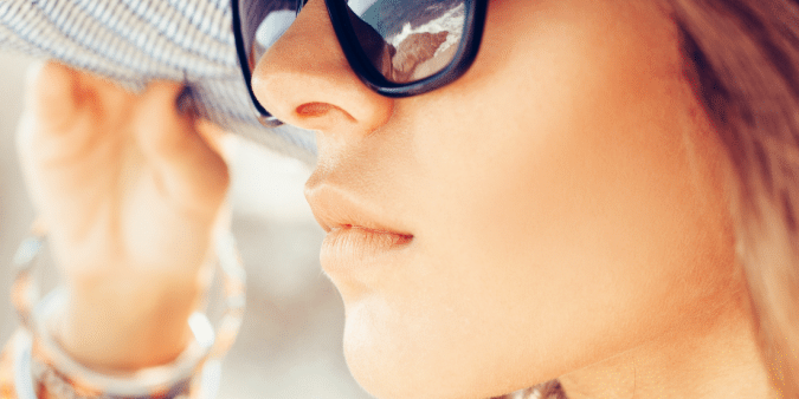 A woman wearing large black sunglasses and a wide-brimmed hat looks into the distance. She holds the brim of her hat with one hand, and colorful bracelets adorn her wrist. The image is bright and summery.