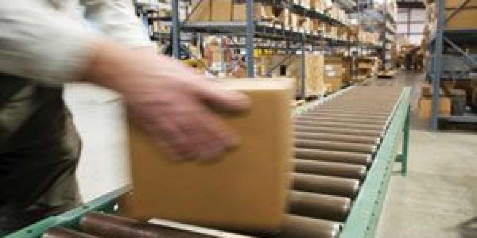 A person places a cardboard box on a conveyor belt in a warehouse with shelves full of boxes and packages in the background.