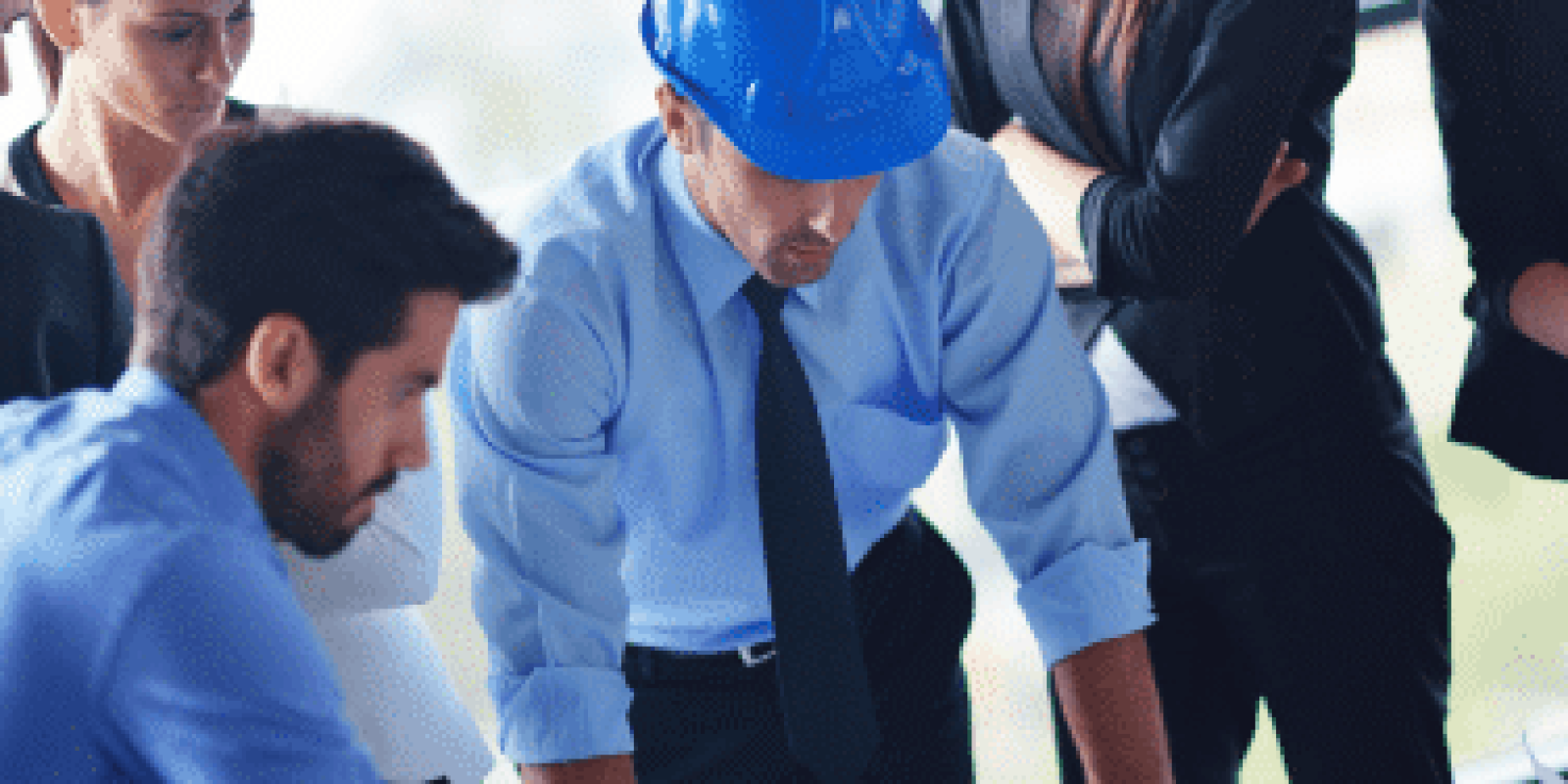 A group of business professionals and an engineer wearing a blue hard hat are gathered around a table, closely examining blueprints or architectural plans in a bright office setting.