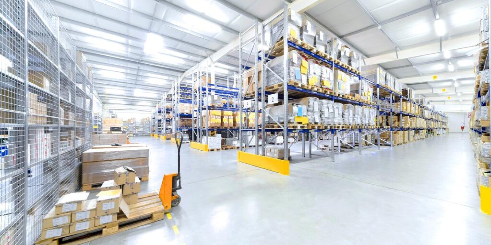 Spacious warehouse with high shelves filled with boxes and goods. Pallets and a hand pallet truck are visible on the polished concrete floor under bright overhead lighting.