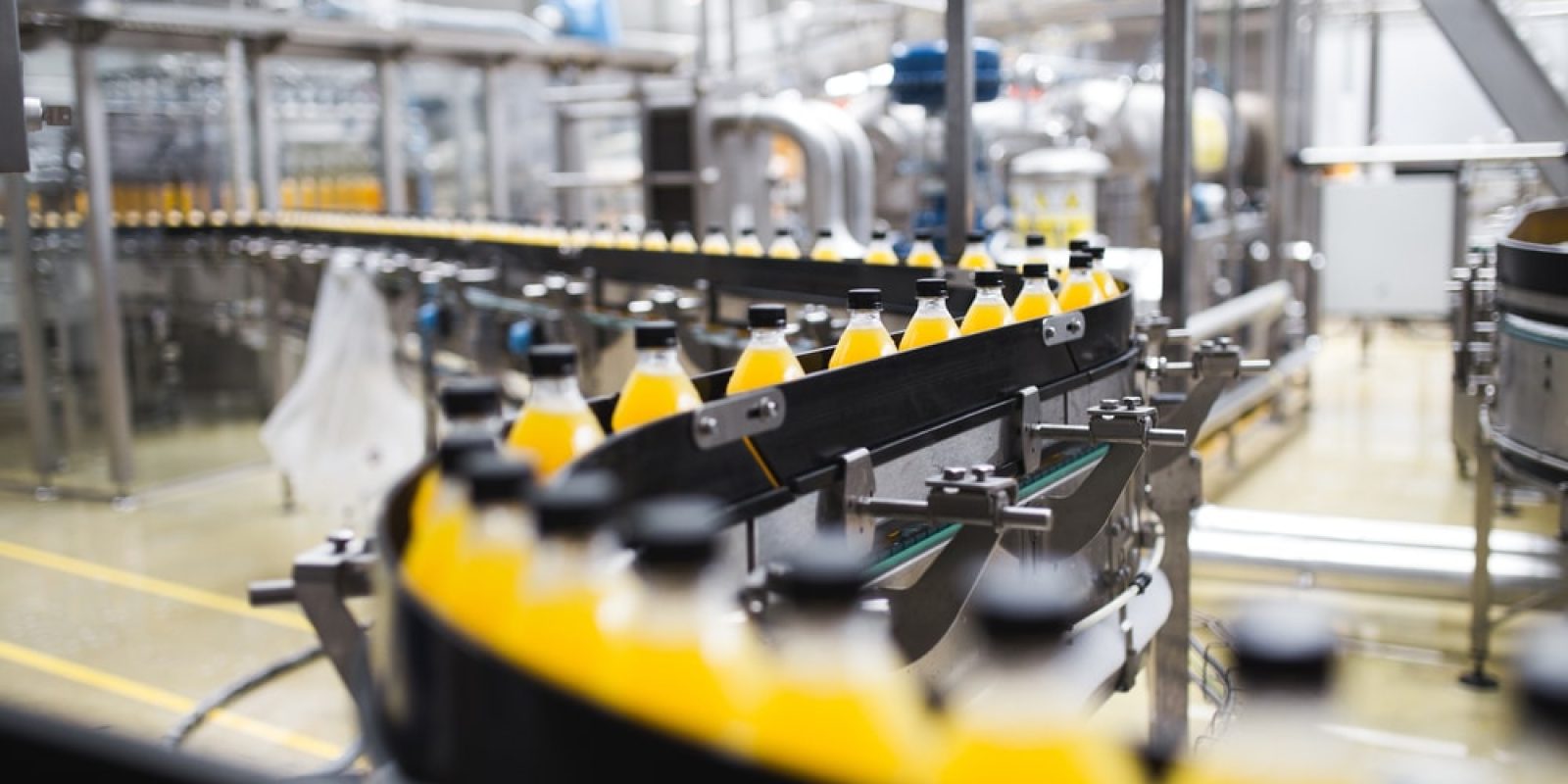 A conveyor belt carries rows of plastic bottles filled with orange liquid through a modern beverage factory, where automation strategies and stainless steel machinery ensure efficient production.
