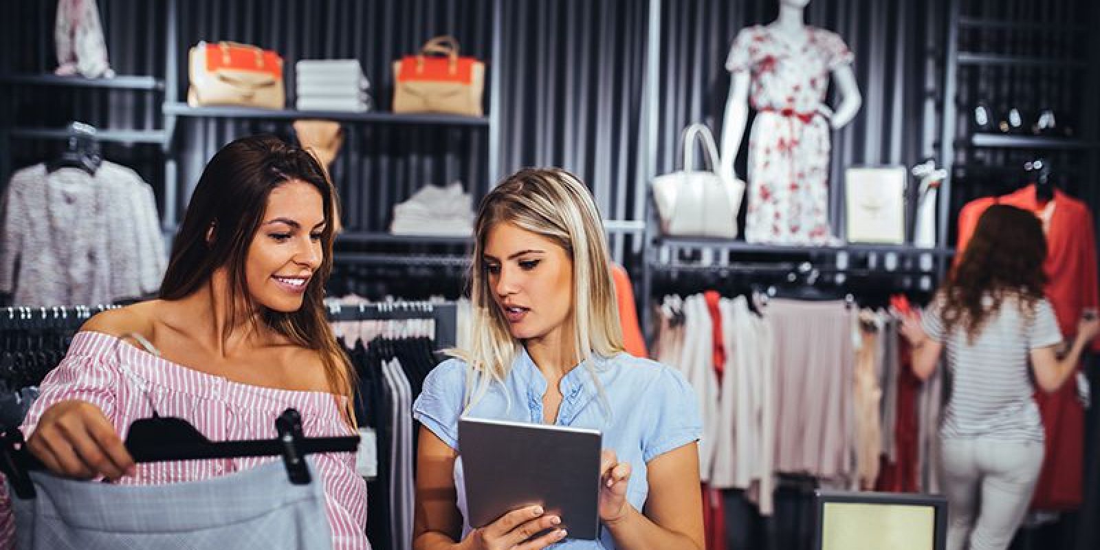 Two women are shopping for clothes in a store; one is holding a garment while the other shows her something on a tablet. Clothing and handbags are displayed in the background, with two more people browsing.