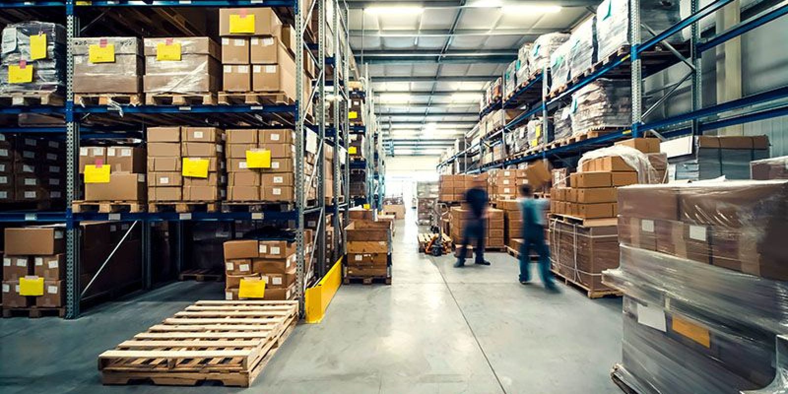 Large warehouse interior with shelves stacked high for optimal warehouse space utilization. Two blurred workers move along the aisles, and wooden pallets are visible on the floor. The brightly lit area ensures efficient operations.