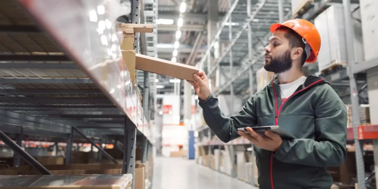 A man wearing an orange hard hat and a green jacket stands in a warehouse, holding a tablet and placing a box on a shelf. The warehouse has tall shelves stocked with various items.